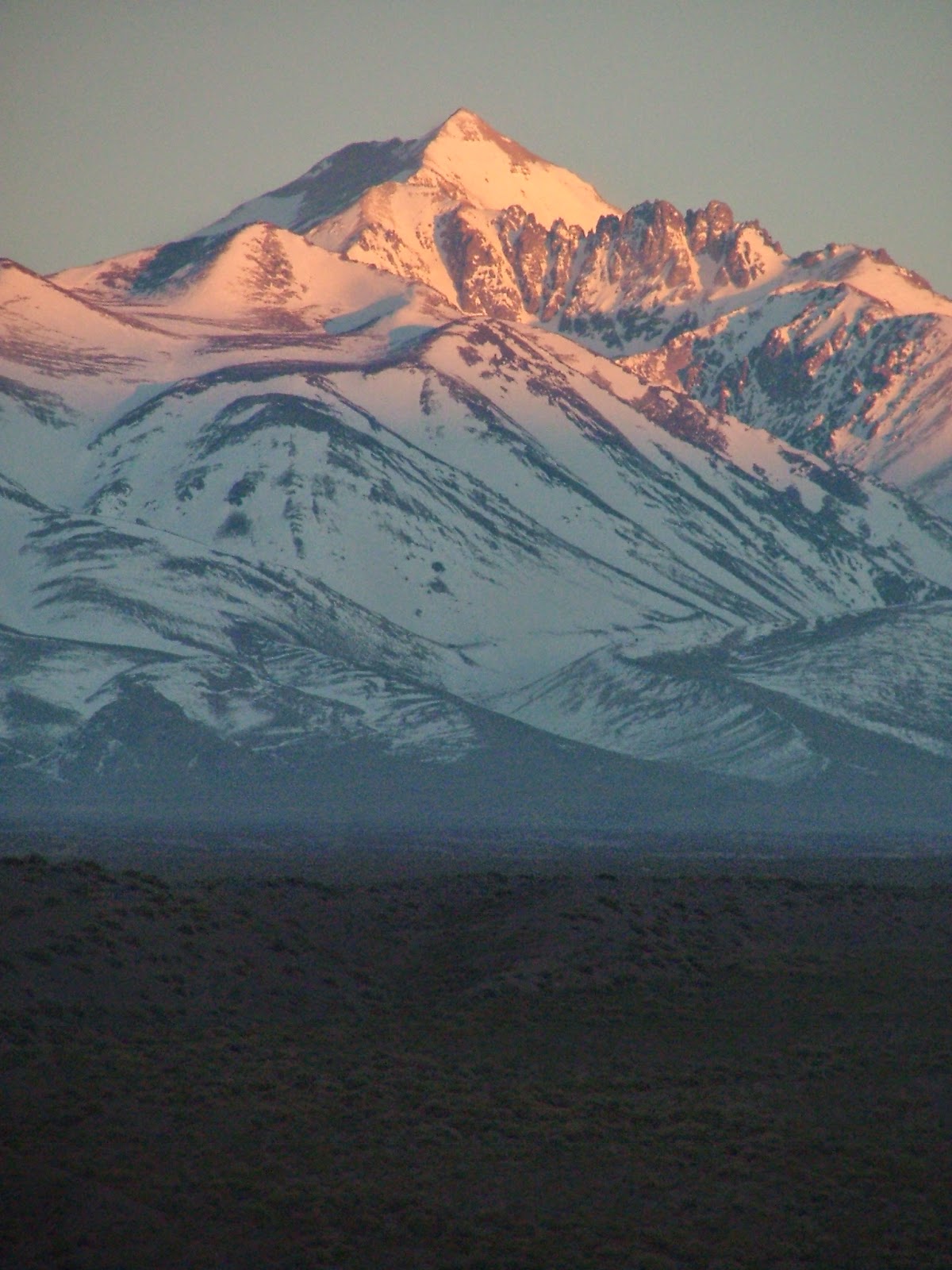 Cadena montañosa nevada iluminada por luz cálida al atardecer con matorrales oscuros en primer plano, tonos naranjas y azules, atmósfera serena.