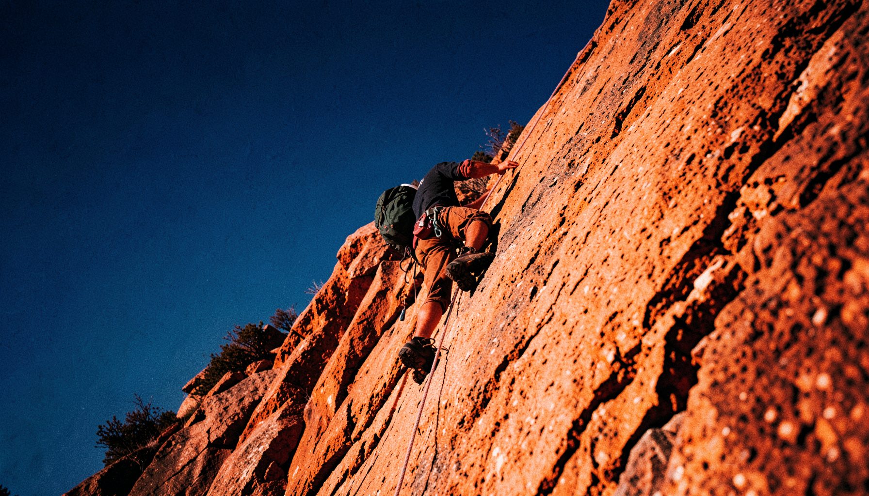 Escalador ascendiendo una pared de roca anaranjada bajo luz solar intensa, con cuerda visible y cielo azul profundo