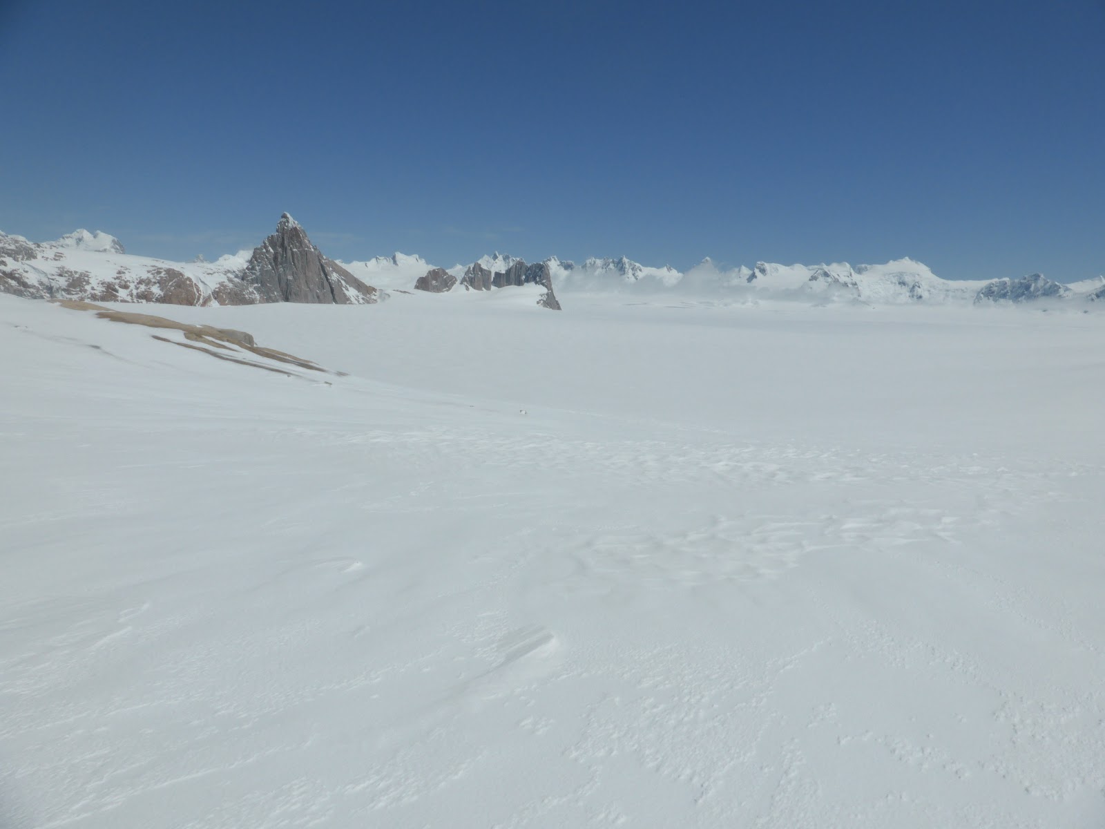 Vasto campo de nieve con picos rocosos y dentados al fondo bajo un cielo azul claro, tonos blancos y azules, atmósfera expansiva y serena.