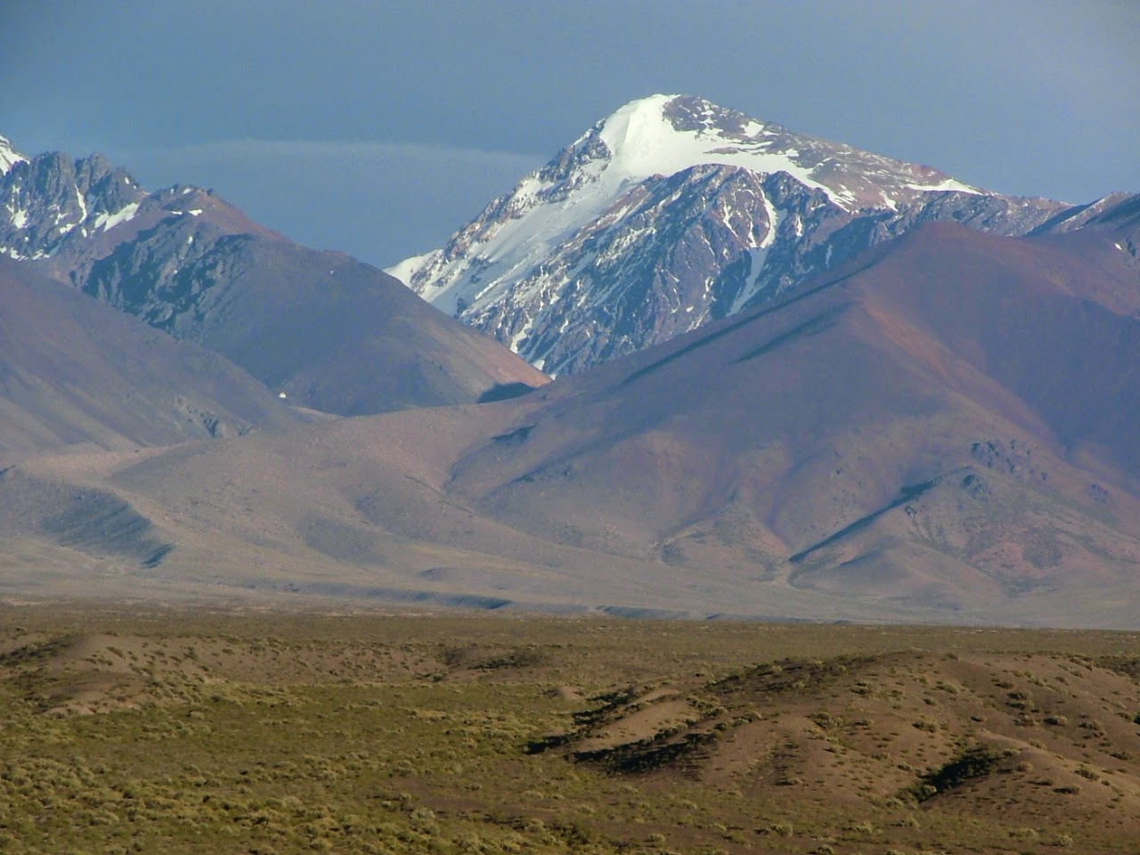 Pico nevado a la distancia detrás de estribaciones marrones y púrpuras, con llanura de arbustos en primer plano bajo cielo claro, tonos suaves y atmósfera serena