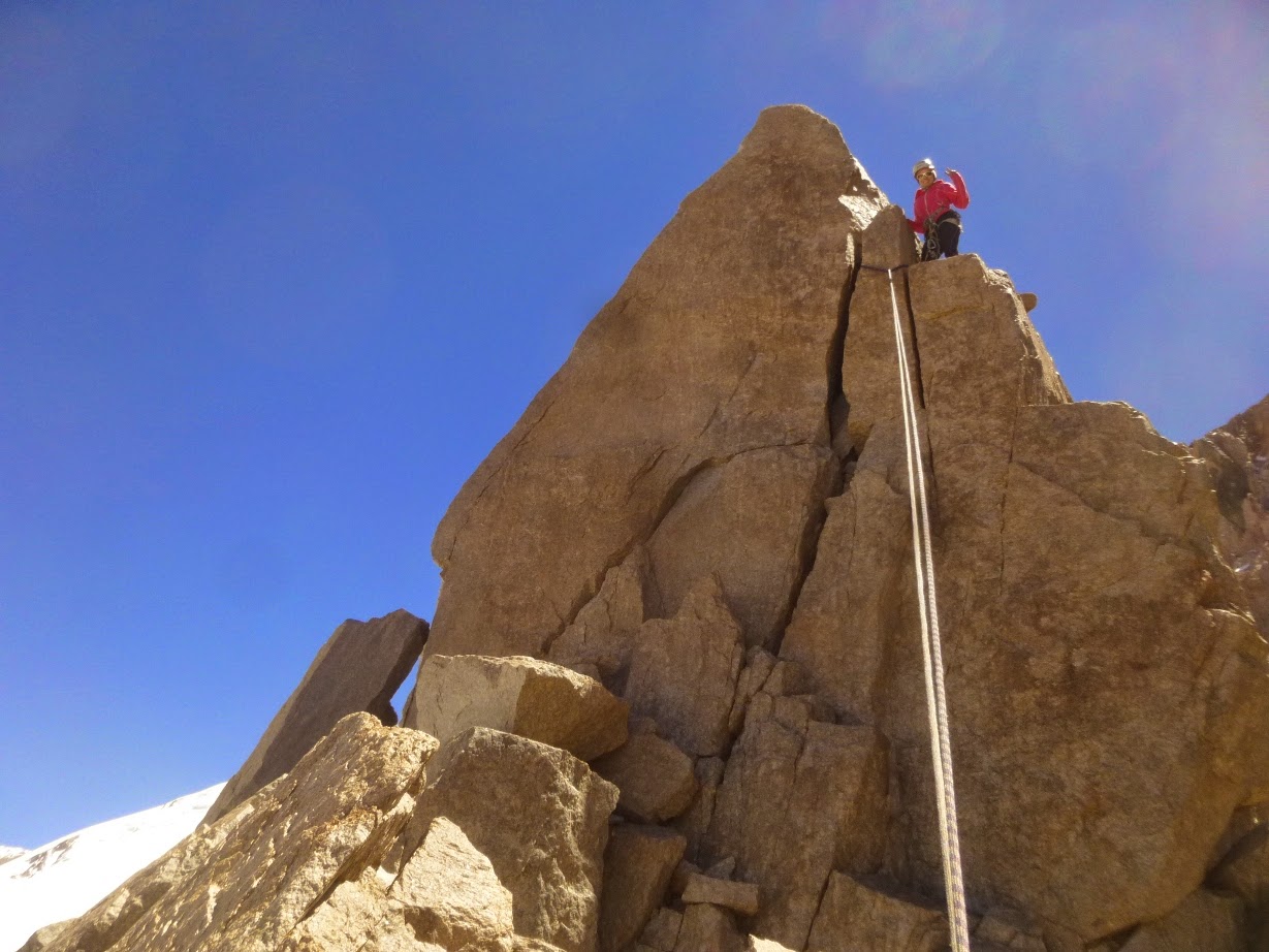 Escalador con chaqueta roja sobre una aguja de roca afilada, con cuerda colgando y un glaciar nevado debajo